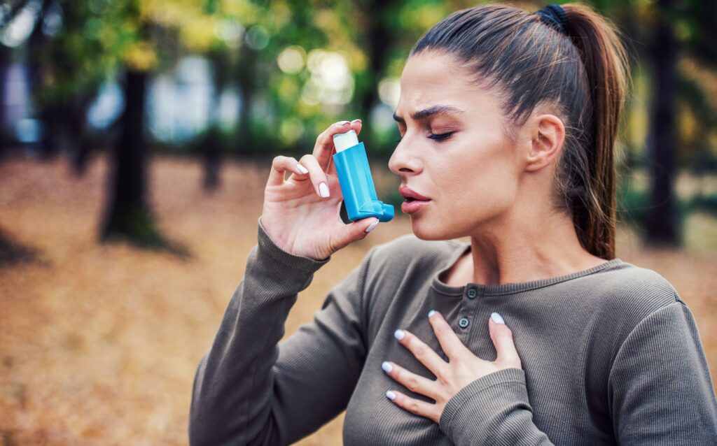 Woman in brown shirt outside holding chest and inhaler looking pained